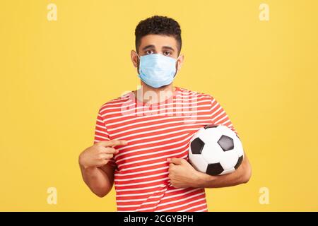 Jeune barbu dans un masque de sécurité et un t-shirt rouge pointant le doigt sur le ballon de football sur sa main avec une expression triste, quarantaine du coronavirus. Studio intérieur Banque D'Images