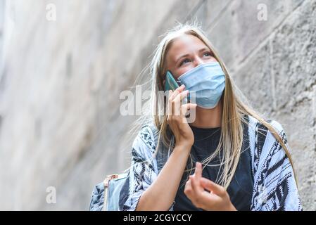 Portrait d'une belle femme blonde caucasienne utilisant un masque bleu parlant par téléphone près d'un bâtiment ancien. Copier l'espace sur la gauche Banque D'Images