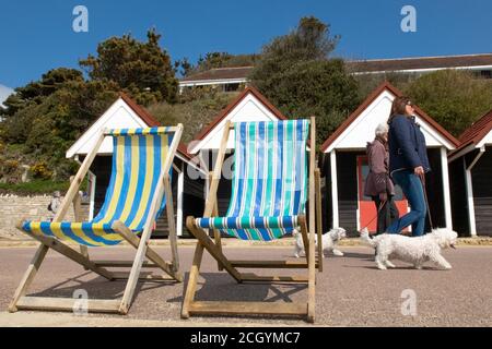 Deux femmes marchent leurs chiens sur la promenade en passant devant des cabanes de plage et des chaises longues à Alum Chine à Bournemouth, Dorset, le matin d'avril. 27 avril 201 Banque D'Images