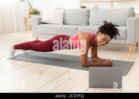 Femme noire faisant de l'exercice de planche à l'ordinateur portable pendant l'entraînement à l'intérieur Banque D'Images