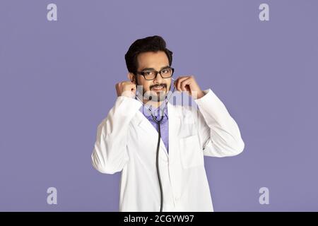Portrait d'un médecin indien joyeux en blouse de laboratoire avec stéthoscope sur fond violet Banque D'Images