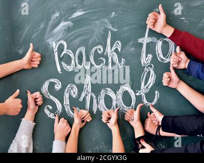 Mains des élèves montrent Thumbs up sur l'arrière-plan de l'inscription retour à l'école à la craie sur un tableau noir de l'école. L'éducation de l'équipe à l'école. Banque D'Images
