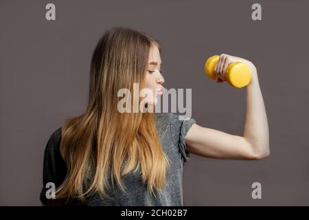 Belle jeune femme aux cheveux blonds amples portant un t-shirt gris tenant une haltère jaune. Prise de vue en studio. Il est temps de visiter le gim Banque D'Images