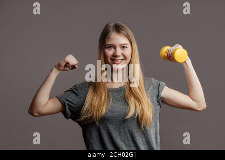 Belle jeune femme de fitness avec des cheveux blonds amples portant un t-shirt gris tenant une haltère jaune. Prise de vue en studio. Banque D'Images