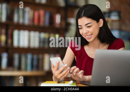 Belle femme asiatique utilisant un smartphone tout en travaillant sur un ordinateur portable En cafétéria Banque D'Images