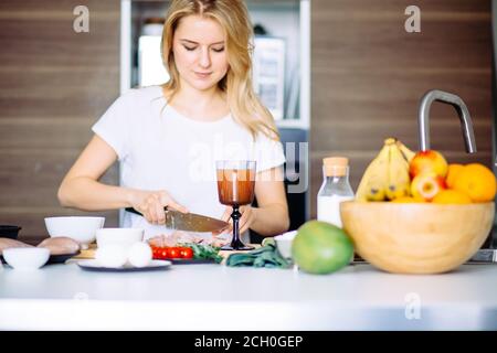Femme au foyer agréable couper la viande sur une table de cuisine. Fruits et légumes colorés, des œufs, de la farine et de smoothie sont placés dans la ligne attendant leur t Banque D'Images