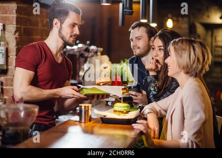 Groupe d'amis souriants heureux commandant de la nourriture par le barkeeper au comptoir dans un restaurant moderne avec style loft, briques et tuyaux, intérieur, Banque D'Images
