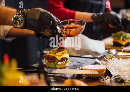 Processus de cuisson des hamburgers. Vue sur les mains du chef avec des gants noirs avec cheeseburger préparé, variété de garnitures et ingridients sur le bureau en bois. Banque D'Images