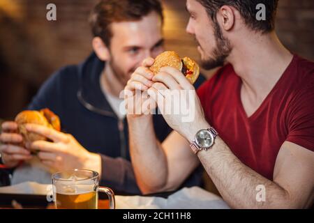 Des amis qui dégusterent de la bière et de délicieux hamburgers lors de la soirée. Le concept de restauration rapide. Délicieux sandwich hamburger malsain dans les mains, prêt à manger. Banque D'Images