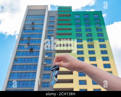 Femme main tenant les clés du doigt à la nouvelle maison. Une main tient les clés du nouvel appartement sur le fond du nouveau bâtiment. Concept immobilier. Banque D'Images