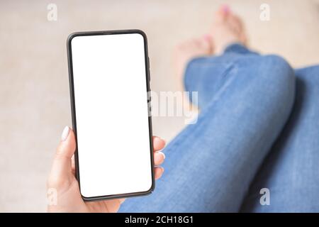 Image de maquette des mains de femme tenant un téléphone portable avec écran vide. La femme est assise sur le canapé avec ses jambes étirées. Pho mobile Mockup Banque D'Images