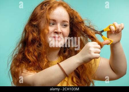 Redhaired femme coupant ses cheveux secs endommagés avec des ciseaux isolés sur fond bleu. Traitement de réparation de cheveux Cure Banque D'Images