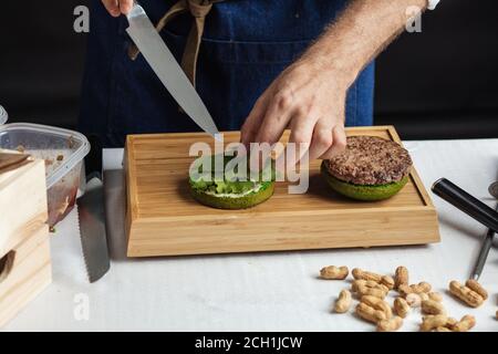 Méconnaissable Male Chef prépare yummy fresh burger avec green bun, épicé sause, viande et fromage. Fermer jusqu'ofcooking concept hamburger. Banque D'Images