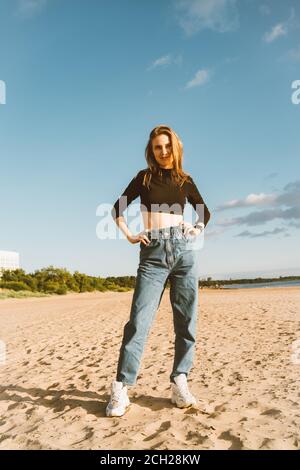 Pleine longueur belle femme debout sur la plage au coucher du soleil en soirée. Jolie femme avec de longs cheveux bruns dans des vêtements décontractés, mains sur les hanches en regardant loin Banque D'Images