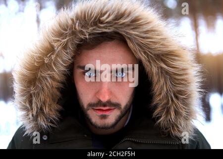 Beau homme sérieux et brutal barbu jeune caucasien debout à l'extérieur de la capuche en fourrure dans la forêt de saison d'hiver. Un sympathique gars européen qui marche en neige Banque D'Images