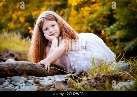 Mignon petite fille de gingembre en robe blanche s'appuie sur un morceau de bois, posant pour une photo Banque D'Images