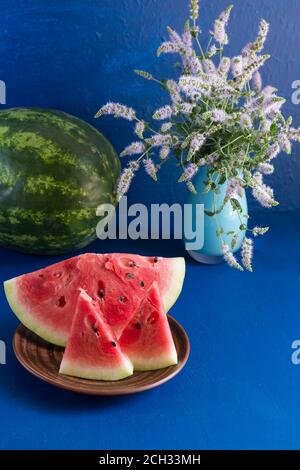 Trois tranches de pastèque fraîche mûre sont dans une assiette en argile sur fond bleu foncé. Theres un melon d'eau entier dans le fond. Dans un vase, un bouquet Banque D'Images