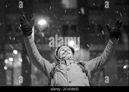 Homme caucasien mature barbu en veste d'hiver debout sous la neige avec les mains soulevées au ciel et regardant vers le haut. Sépia Banque D'Images