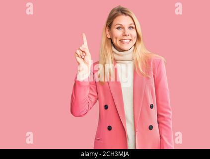 Jeune belle femme blonde portant des vêtements élégants montrant et pointant vers le haut avec le doigt numéro un tout en souriant confiant et heureux. Banque D'Images