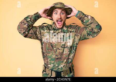 Jeune homme caucasien portant l'uniforme militaire de camouflage fou et effrayé avec les mains sur la tête, peur et surpris de choc avec la bouche ouverte Banque D'Images