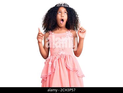 Enfant afro-américain aux cheveux bouclés portant la couronne de princesse stupéfait et surpris regardant vers le haut et pointant avec les doigts et les bras relevés. Banque D'Images
