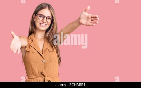 Jeune belle femme blonde portant des vêtements décontractés et des lunettes regardant l'appareil photo sourire avec les bras ouverts pour le câlin. Expression gaie embrassant ha Banque D'Images