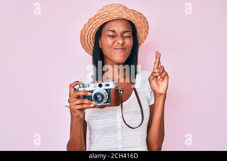 Jeune femme afro-américaine portant un chapeau d'été tenant un appareil photo vintage gestante doigt croisé souriant avec espoir et les yeux fermés. Chance et superstiti Banque D'Images