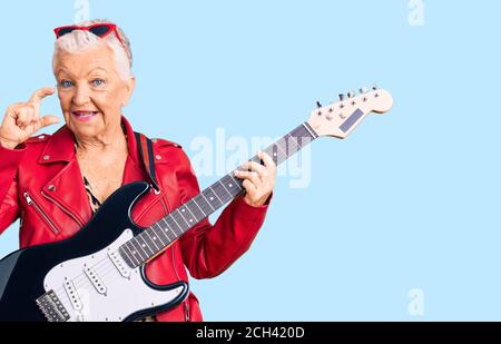 Femme senior belle avec les yeux bleus et les cheveux gris port un look moderne jouant de la guitare électrique souriant et confiant gestante à la main, faire des taches Banque D'Images