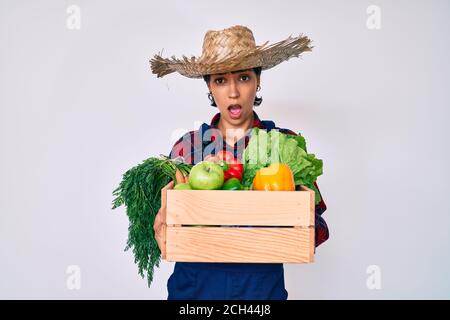 Belle femme brunettte portant des vêtements de fermier tenant des légumes dans le visage de choc, regardant sceptique et sarcastique, surpris par la bouche ouverte Banque D'Images