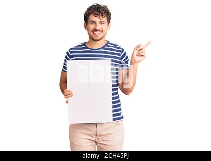 Jeune homme beau avec cheveux bouclés tenant une bannière vide sourire heureux pointant avec la main et le doigt sur le côté Banque D'Images