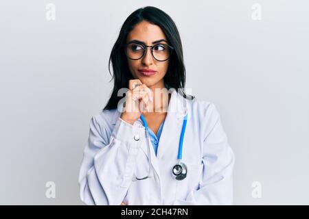 Belle femme hispanique portant l'uniforme de médecin et la pensée de stéthoscope concentrée au sujet du doute avec le doigt sur le menton et regardant vers le haut se demander Banque D'Images