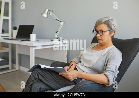 Femme mûre occupée avec des lunettes écrit du texte dans un bloc-notes tout en étant assise dans une chaise à la maison. Femme âgée étudiant à la maison. Banque D'Images