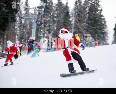 Bukovel, Ukraine - 09 décembre 2018 : homme snowboarder en costume de Père Noël avec barbe blanche tenant le monopied de selfie à la main, freeride sur le snowboard lors d'une compétition de freeride sur une piste de ski enneigée Banque D'Images