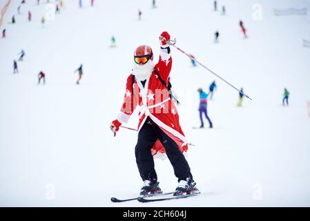 Bukovel, Ukraine - 09 décembre 2018 : homme skieur à Santa Clause costume avec barbe blanche dans des lunettes et un casque, ski vers le bas et levant la main avec un bâton de ski sur une pente enneigée très peuplée Banque D'Images