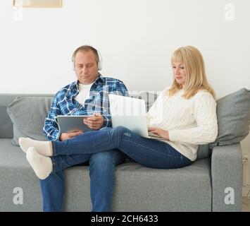 Portrait complet d'un couple d'âge mûr avec addiction numérique à la maison. Beau homme avec une tablette et belle femme d'âge moyen avec un ordinateur portable passer du temps ensemble tout en étant allongé sur un canapé. Banque D'Images