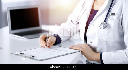 Une femme-médecin inconnue prescrit des médicaments à son patient, à l'aide d'un presse-papiers, tout en étant assise au bureau dans son cabinet ensoleillé. Femme Banque D'Images