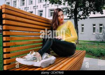 Cette jolie fille aux cheveux longs et foncés porte un chandail jaune vif, des leggings foncés brillants et serrés et des baskets dorées. Elle est assise sur un banc en bois Banque D'Images