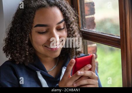 Belle race mixte afro-américaine fille adolescente jeune femme heureux sourire assis près d'une fenêtre à l'aide d'un téléphone portable Banque D'Images