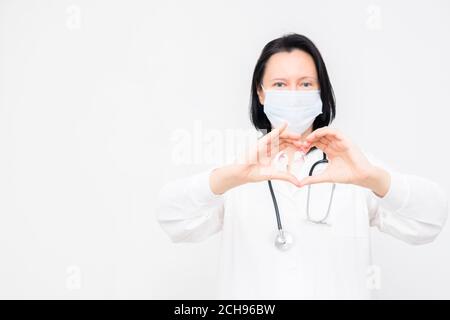 Femme médecin dans le masque médical montre le symbole de coeur sur fond blanc avec espace de copie. Banque D'Images