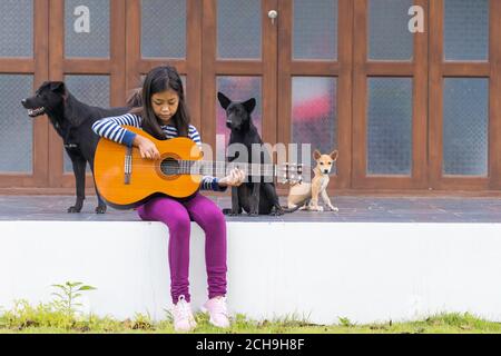 Une mignonne écolière asiatique est heureuse de jouer de la guitare avec son chien à la maison. Banque D'Images