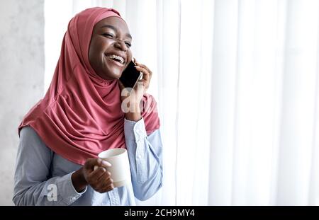 Joyeuse femme musulmane noire qui bume du café et parle au téléphone portable Près de la fenêtre Banque D'Images