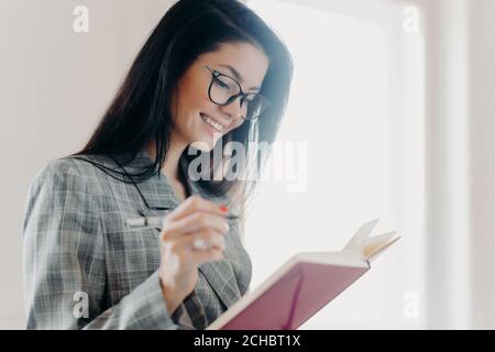 Prise de vue latérale de la femme brune agréable à regarder a l'expression heureuse fait des notes dans le planificateur, fait le travail à distance, la planification des creats, porte des lunettes optiques Banque D'Images