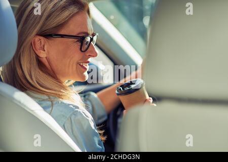 Vue arrière d'une belle femme d'affaires d'âge moyen portant des lunettes de vue assise derrière le volant dans une voiture, buvant du café et souriant Banque D'Images