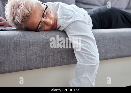 une enseignante fatiguée dorme sur un canapé pendant la leçon en ligne, elle est épuisée pour parler et vérifier ses travaux Banque D'Images