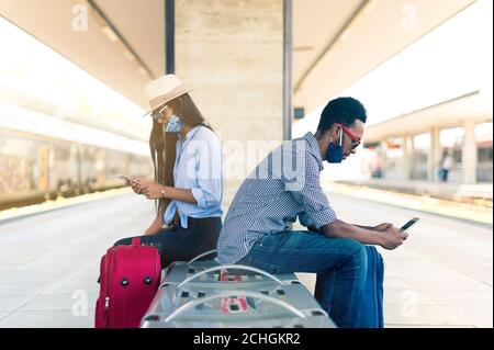 Couple assis sur un banc entre deux chemins de fer - jeunes Avec masque facial utilisant des téléphones intelligents mobiles - Voyage pandémique et concept de style de vie Banque D'Images