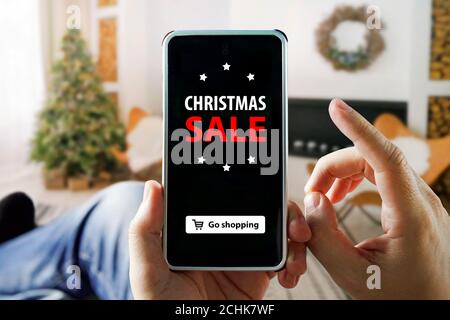 Noël shopping en ligne avec téléphone. Man est assis dans un appartement et fait des achats par un smartphone dans un magasin en ligne. Arbre de Noël, cadeaux Banque D'Images