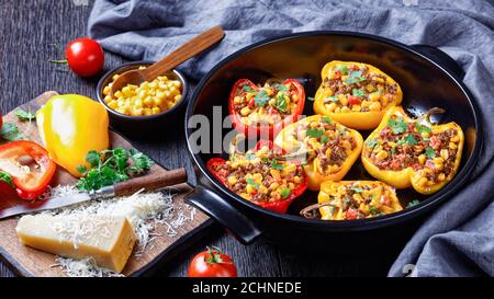 poivrons farcis au bœuf haché, au maïs et au fromage dans un plat de cuisson noir sur une table en bois avec des ingrédients sur une planche à découper, vue horizontale f Banque D'Images