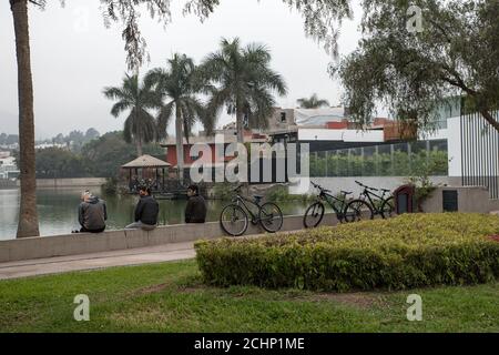 Jeunes hommes portant un masque facial assis près d'un lac avec leurs vélos sur le côté, Lima, Pérou. Banque D'Images