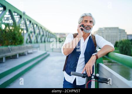 Portrait d'un homme mature avec un scooter électrique en plein air en ville, à l'aide d'un smartphone. Banque D'Images