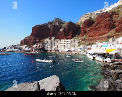 Des bateaux amarrés dans la baie d'Amoudi sous un ciel bleu clair et contre des falaises rouges et des maisons blanches d'Oia, Santorin Banque D'Images